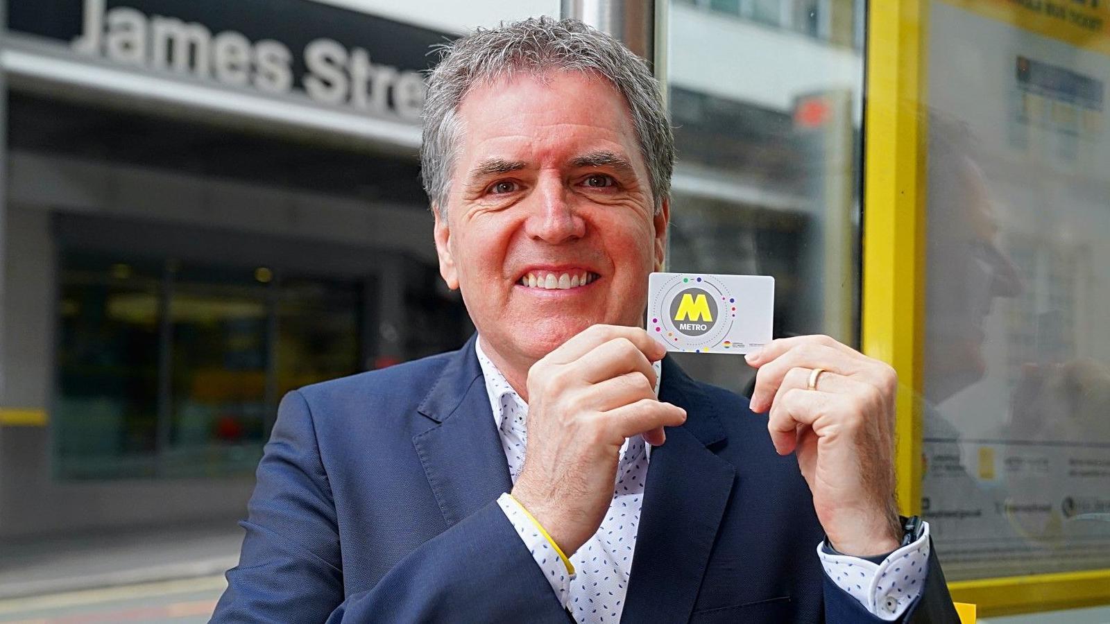 Metro Mayor Steve Rotheram in a blue suit with a white shirt with blue dots. He is holding up a white card with a large letter M on it and smiling at the camera. He is stood at a yellow framed bus stop outside James Street station.
