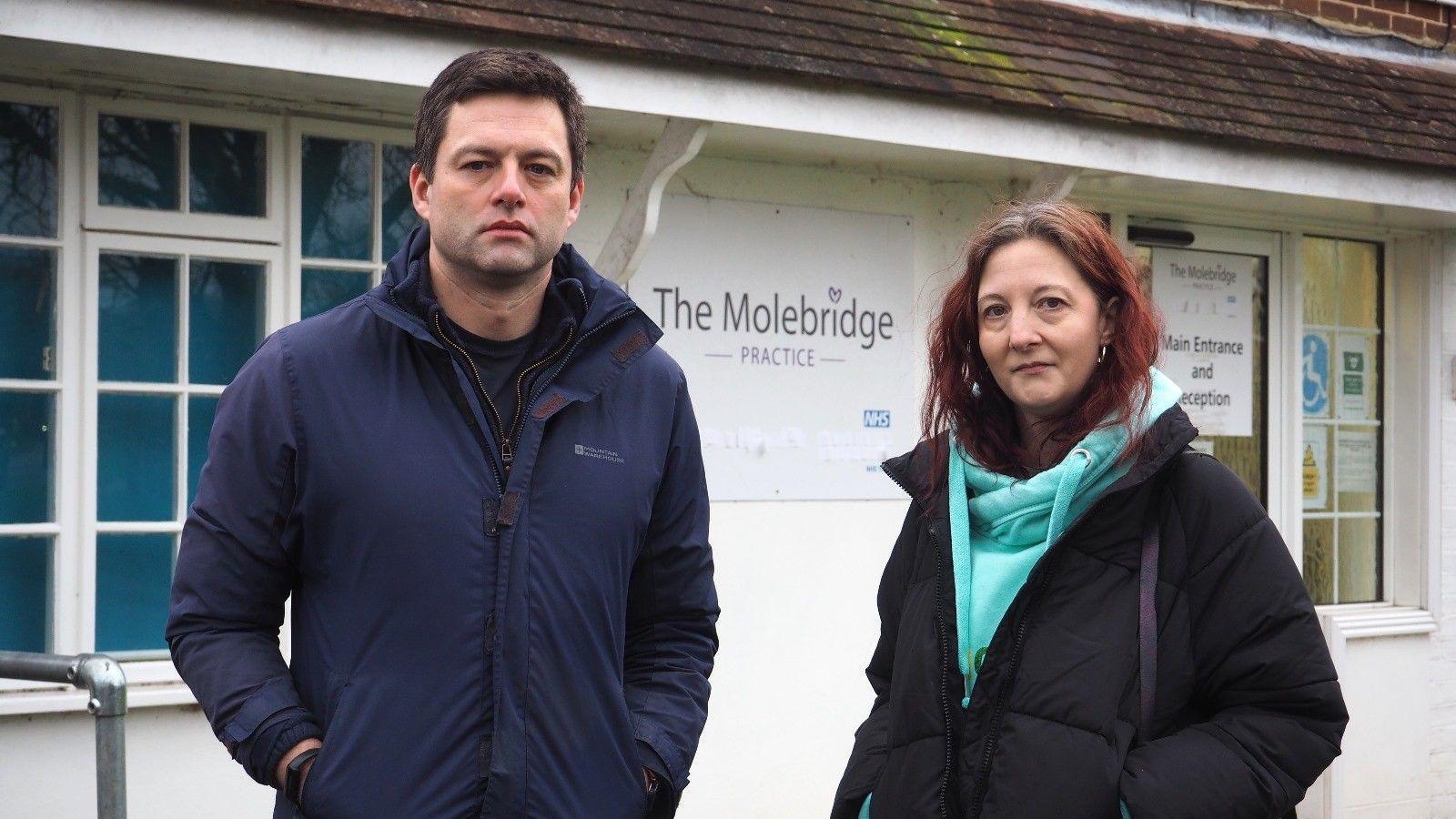 Chris Coghlan MP, left, wearing a navy blue coat, and Caroline Joseph, right, wearing a light blue hoodie and a black coat. They are stood in front of The Molebridge Practice in Fetcham, a white building with a sign reading "The Molebridge Practice" on the front.