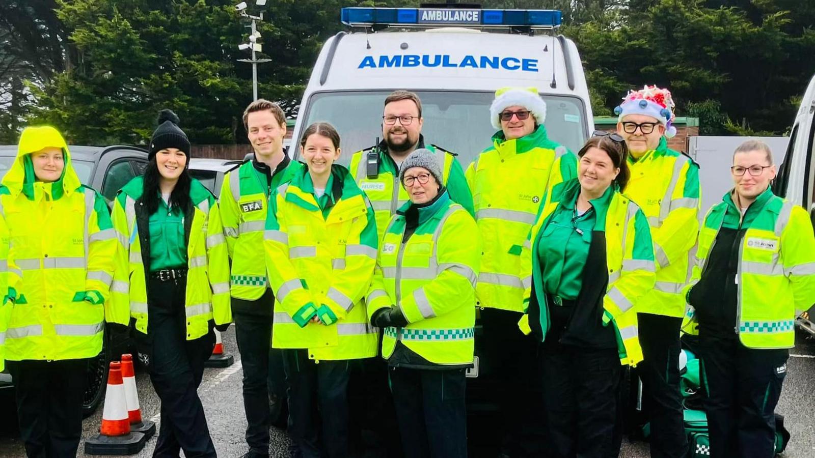 10 o wirfoddolwyr St John yn sefyll o flaen ambiwlans yr elusen. Maen nhw i gyd yn gwisgo siacedi llachar melyn. Mae rhai yn gwisgo hetiau.