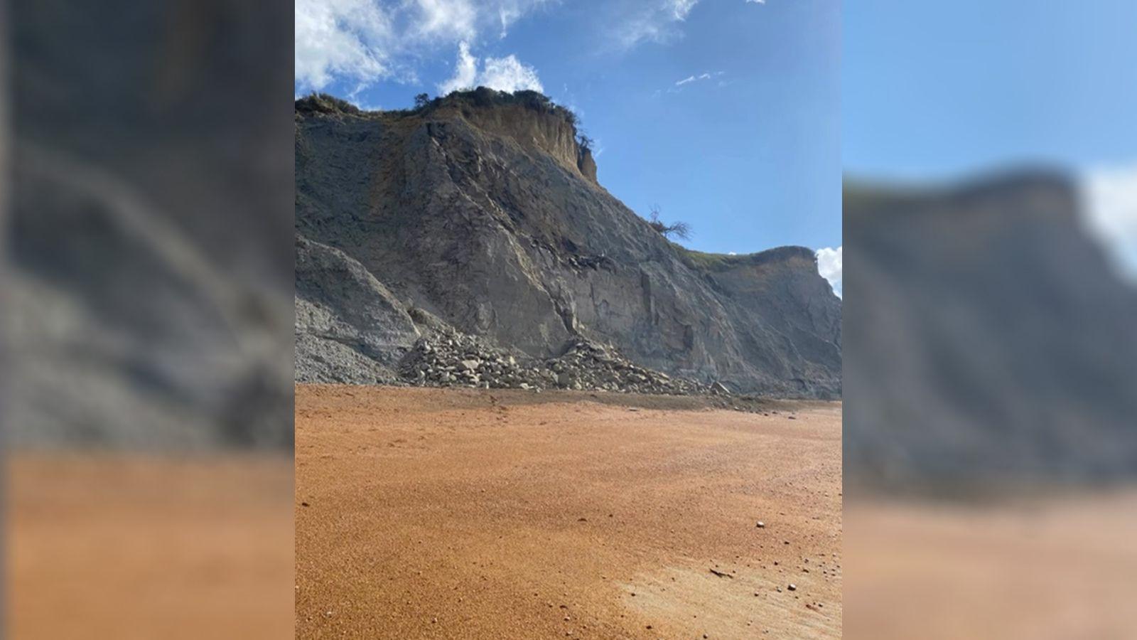 A pile of rocks at the base of a crumbling grey cliff. In the foreground is a sandy coloured beach.