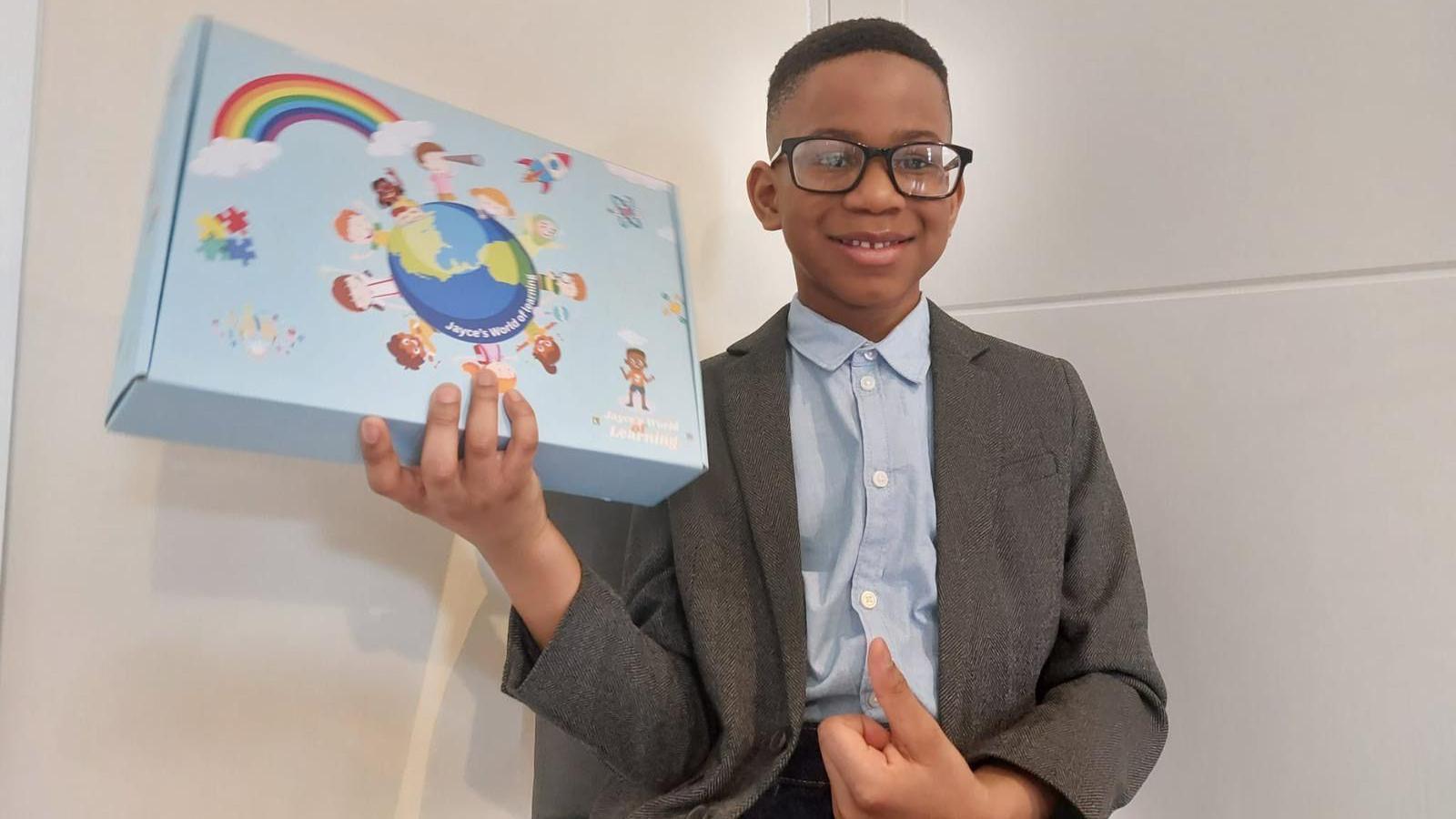 Jayce, a boy with short dark hair and glasses, wearing a grey jacket and blue buttoned-up shirt while holding up a blue box with images of children on.