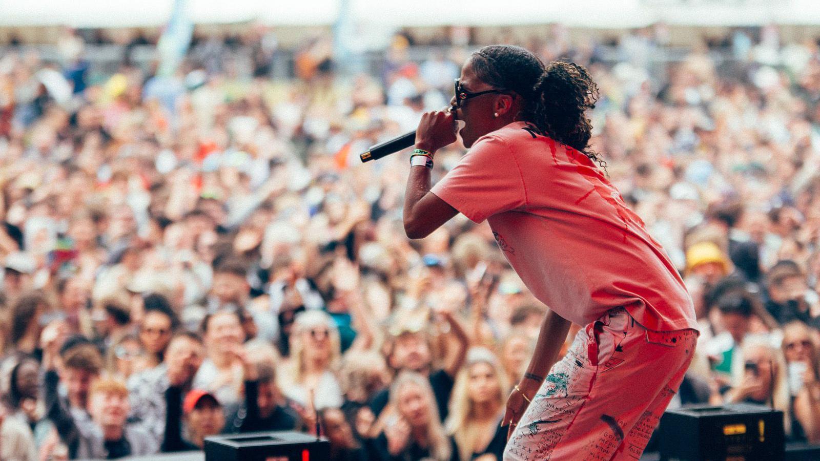 Ms Dynamite, in T-shirt and trousers, is on stage, holding the mic to her mouth. A big crowd is seen behind her with blurred faces.