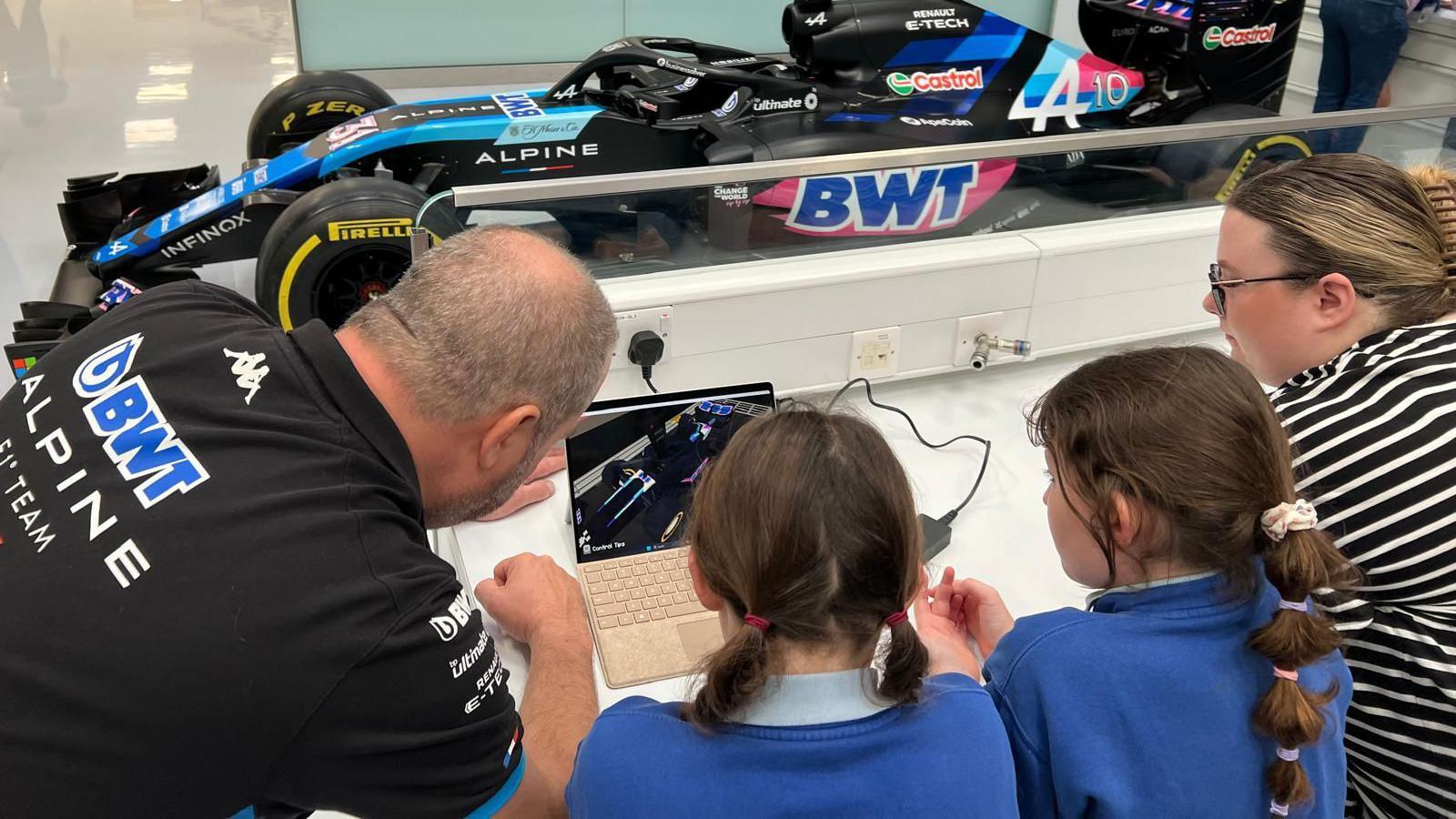 Four people crowded around a laptop screen. The one on the left is a man wearing a black shirt, the middle two are school girls wearing blue sweatshirts and on the right is a woman with a black and white stripy top on. In the background is a pink and blue formula one car.