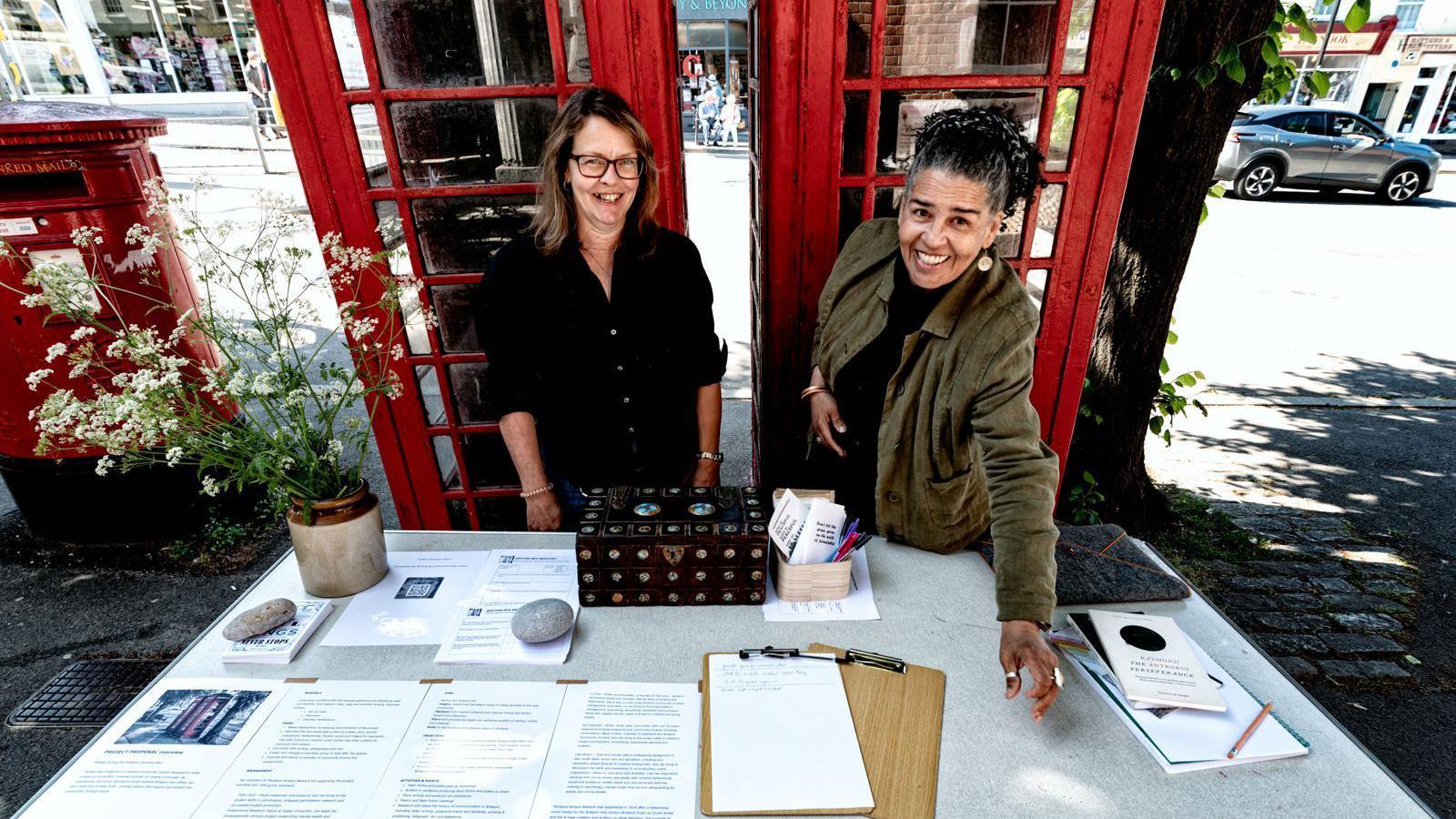 Lisa Wilson and Kim Squirrell standing behind a table next to the two red phone boxes. The table is full of information sheets, clipboards and pens. They are both looking up towards the camera and laughing.
