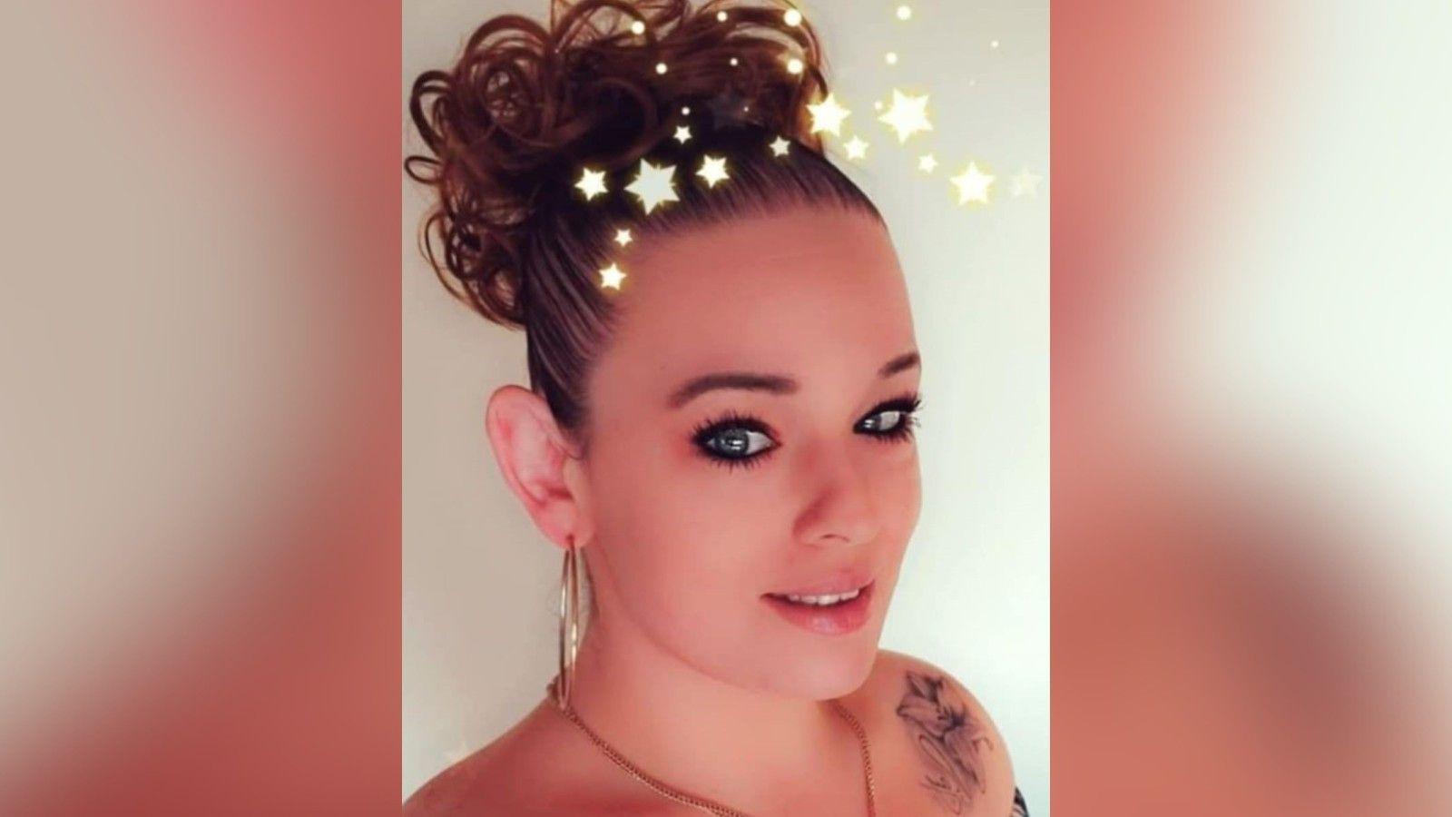 Tammy Morris pictured with her hair in a slick high brown bun and flower tattoo on her right shoulder. She has a large gold, hooped earring in her right ear.
