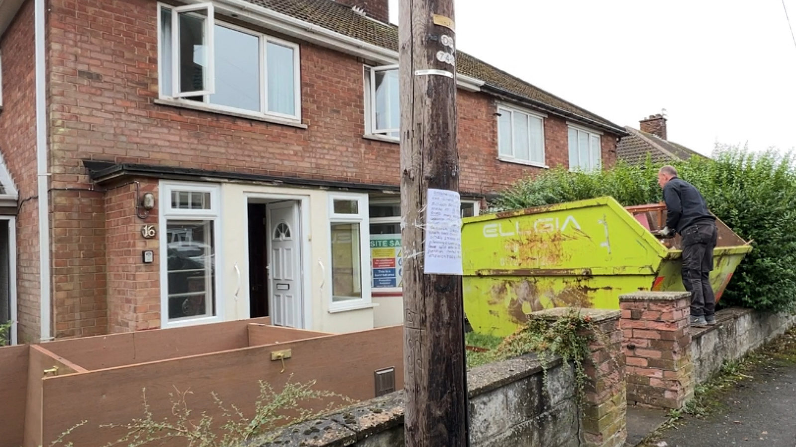 An image of a house, a green skip is towards the right of the frame as a man is picking items from it. The house's white door is open as are the upstairs windows. In front of the property taped to a pole is a handwritten note which lists disagreements with the plans for the house.