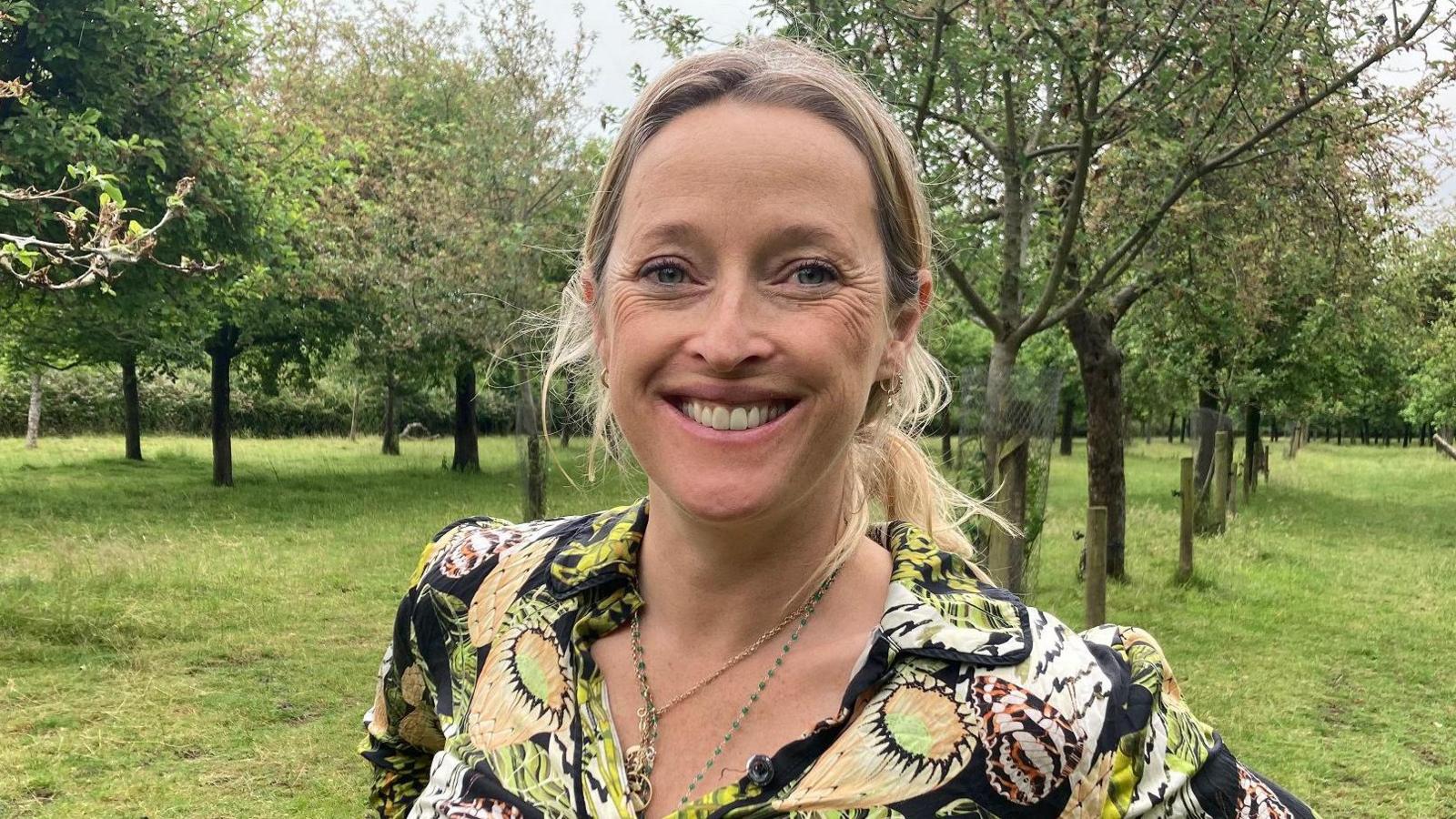 Woman in patterned shirt stands in front of trees in an orchard