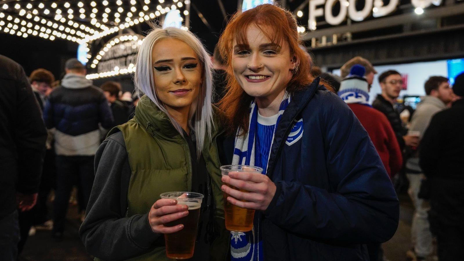 Two women, one with ginger hair and one with white blonde hair, holding pings of beer and smiling at the camera. There's a food stall and some fairy lights behind them, as well as crowds of people.