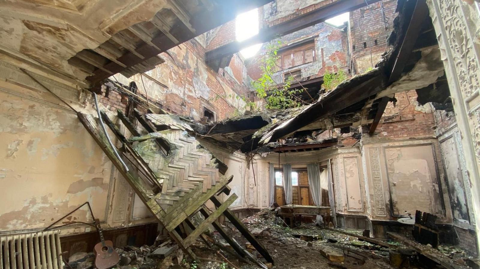 The inside of a room of an old manor house. The ceiling has caved in, a tree is growing through the roof and the ground is covered in debris. A guitar is discarded in one corner.
