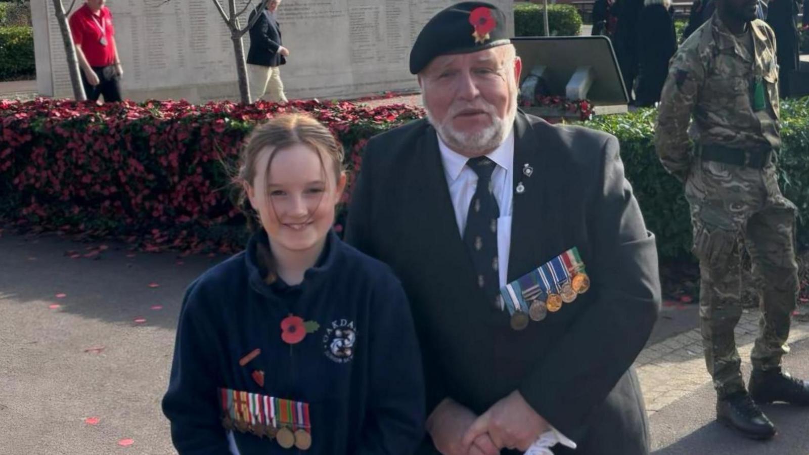 Olivia and her grandfather Terry Clarkson posing for the camera - both are wearing military medals and remembrance poppies. Terry is wearing a military cap, blazer and tie.