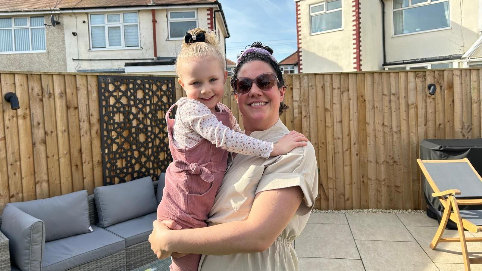 A woman, wearing sunglasses, holds a four-year-old girl in a garden on a sunny day.