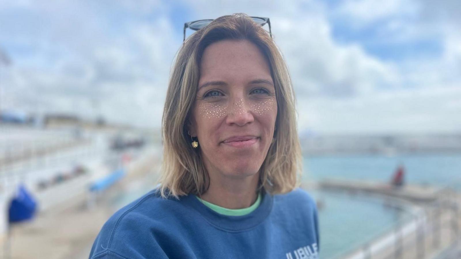 Nicola Murdoch who is wearing a blue jumper and has gold glitter on her face smiles at the camera while standing in front of Penzance's Jubilee Pool, which is blurred in the background.