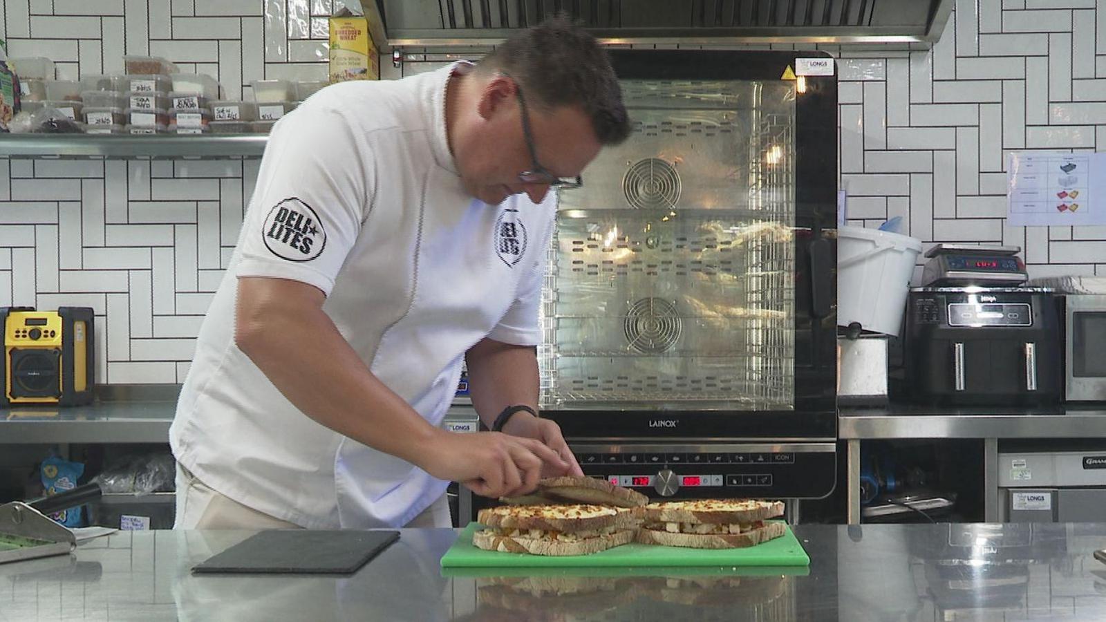 A man with short dark hair and glasses and dressed all in white cuts two sandwiches he has made in half