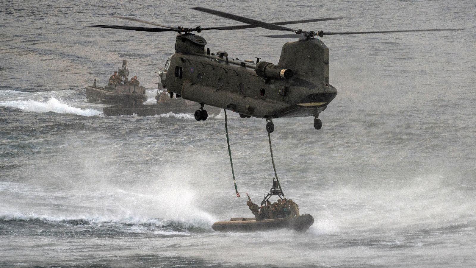 The helicopter is whipping up the water which is causing lots of spray and waves. There are ropes dangling, which are being attached to a dinghy below it.
