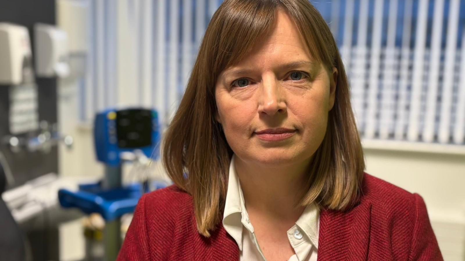 Dr Rees is wearing a cream blouse and red jacket. She has shoulder-length brown hair and is in one of the rooms at the university with clinical equipment in the background.