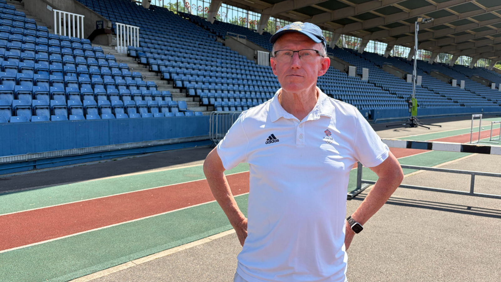 A man stands with his hands on his hips, wearing a white T-shirt, in front of a large athletics stand at a track.