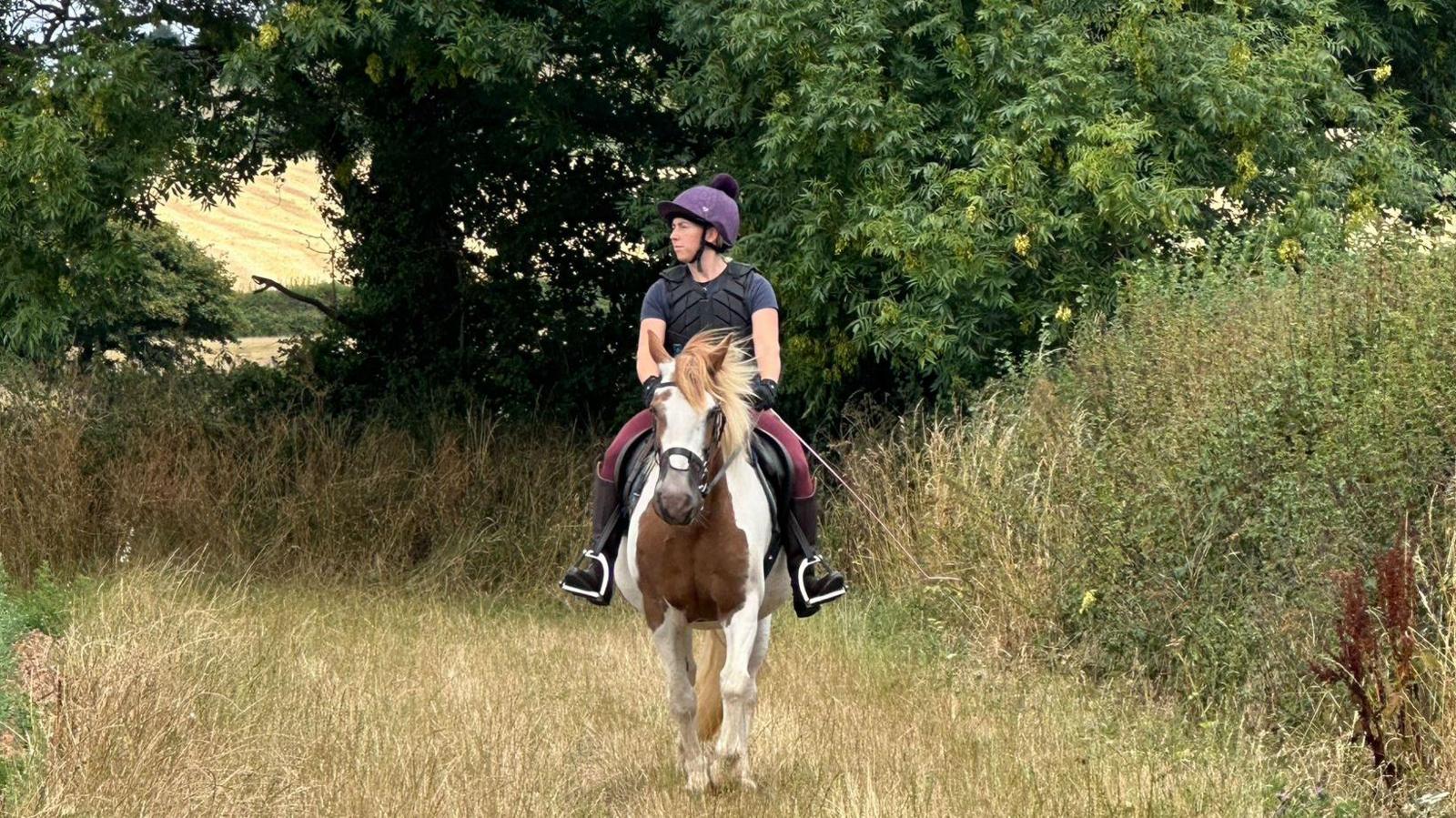 A picture of Laura riding her pony. She is wearing wellies and a purple outfit. She is riding a brown and white horse with blonde hair. She is surrounded by grassland and trees.
