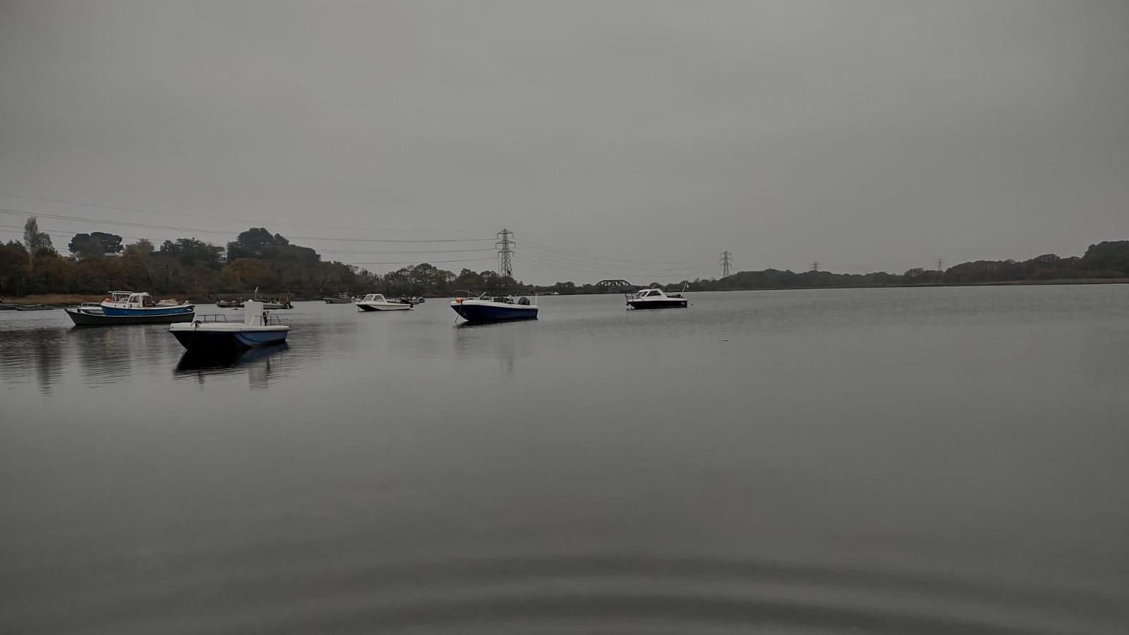 Eight small motorboats are moored in Hamworthy harbour. The water is grey and matches the colour of the sky. On the horizon you can see trees and electricty pylons on a gloomy day.