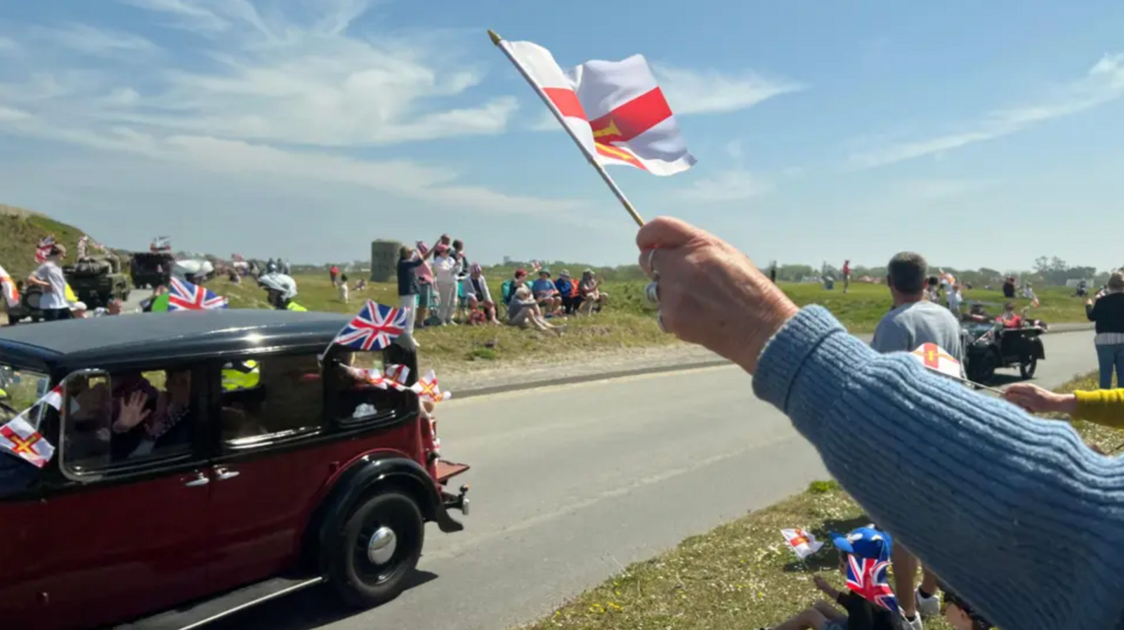 A hand holding a Guernsey flag waving in the air as a WW2-era car drives past, with people lining both of sides of the road celebrating Liberation Day.