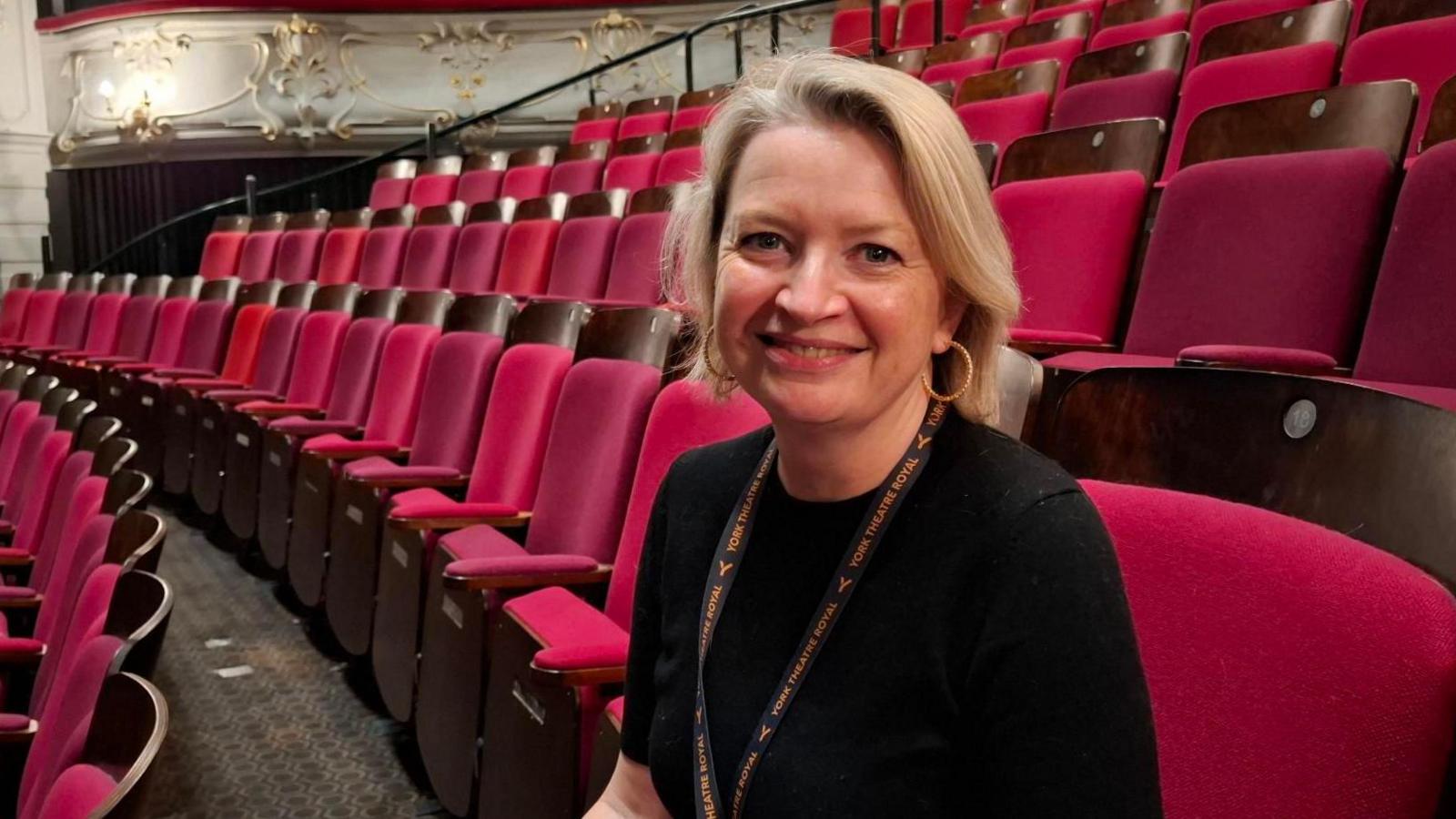 A woman with blonde hair sits in an empty theatre auditorium. She is looking at the camera and smiling. Her lanyard says "York Theatre Royal".