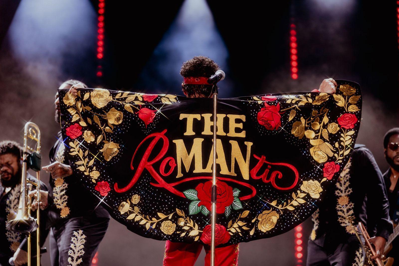 Bruno Mars has his back to the audience on stage at Wembley Stadium. He is wearing a black cape that has red and gold roses on it and the words The Romantic.