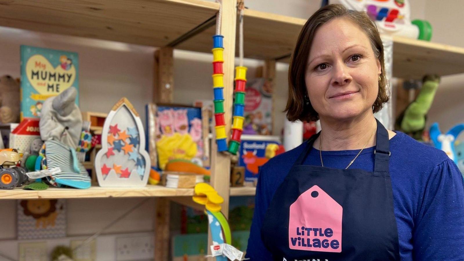 Angela is standing in front of shelves filled with toys. She has a blue t-shirt on and an apron which says Little Village on it