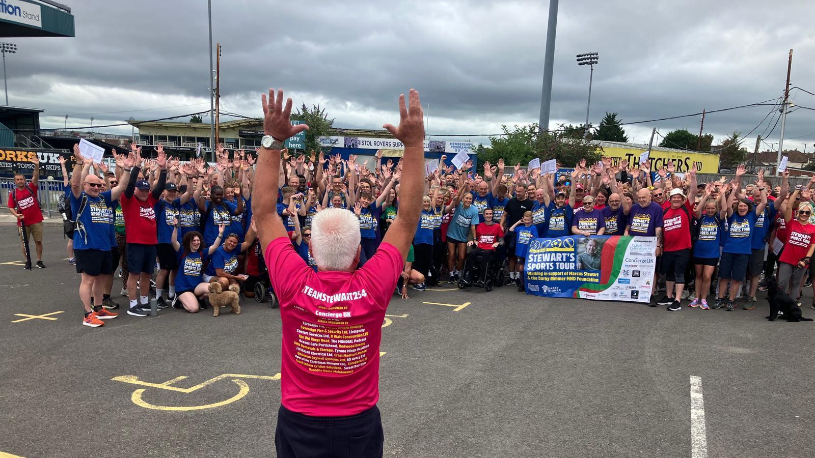 A large crowd of people with their arms in the air standing in front of a man with white hair, who is holding his arms up in the air and wearing a pink shirt. Everyone's t-shirts say 'Stewart's Sports Tour'