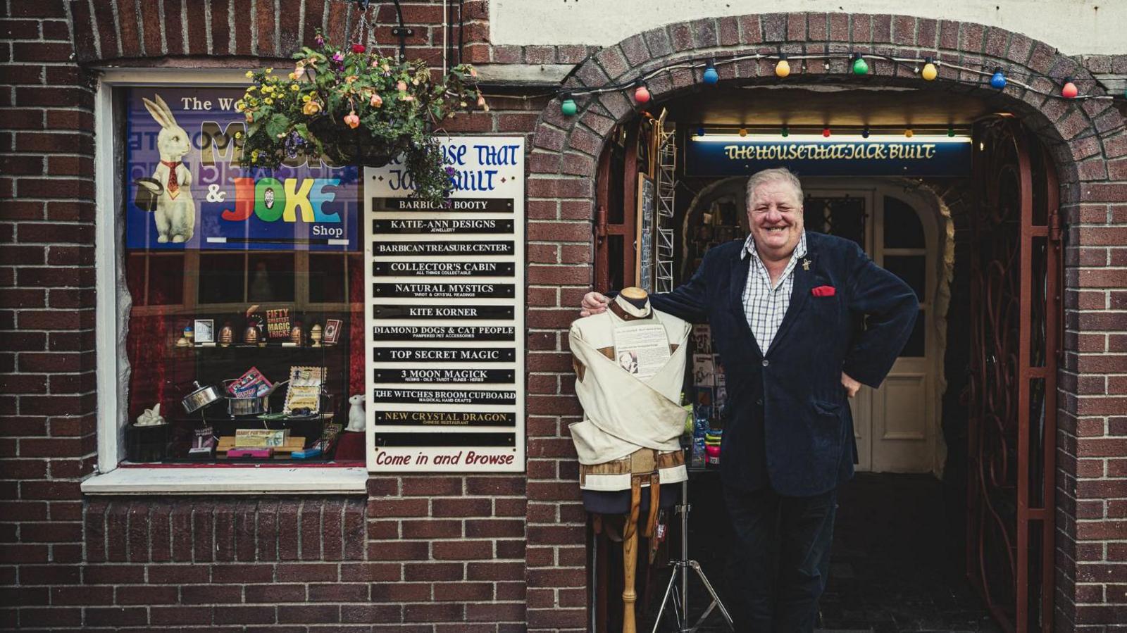 Malcolm Norton stood outside his shop next to a straitjacket, which is on a clothes stand. In the window of the shop there are magic tricks and a sign with a white rabbit holding a hat. 