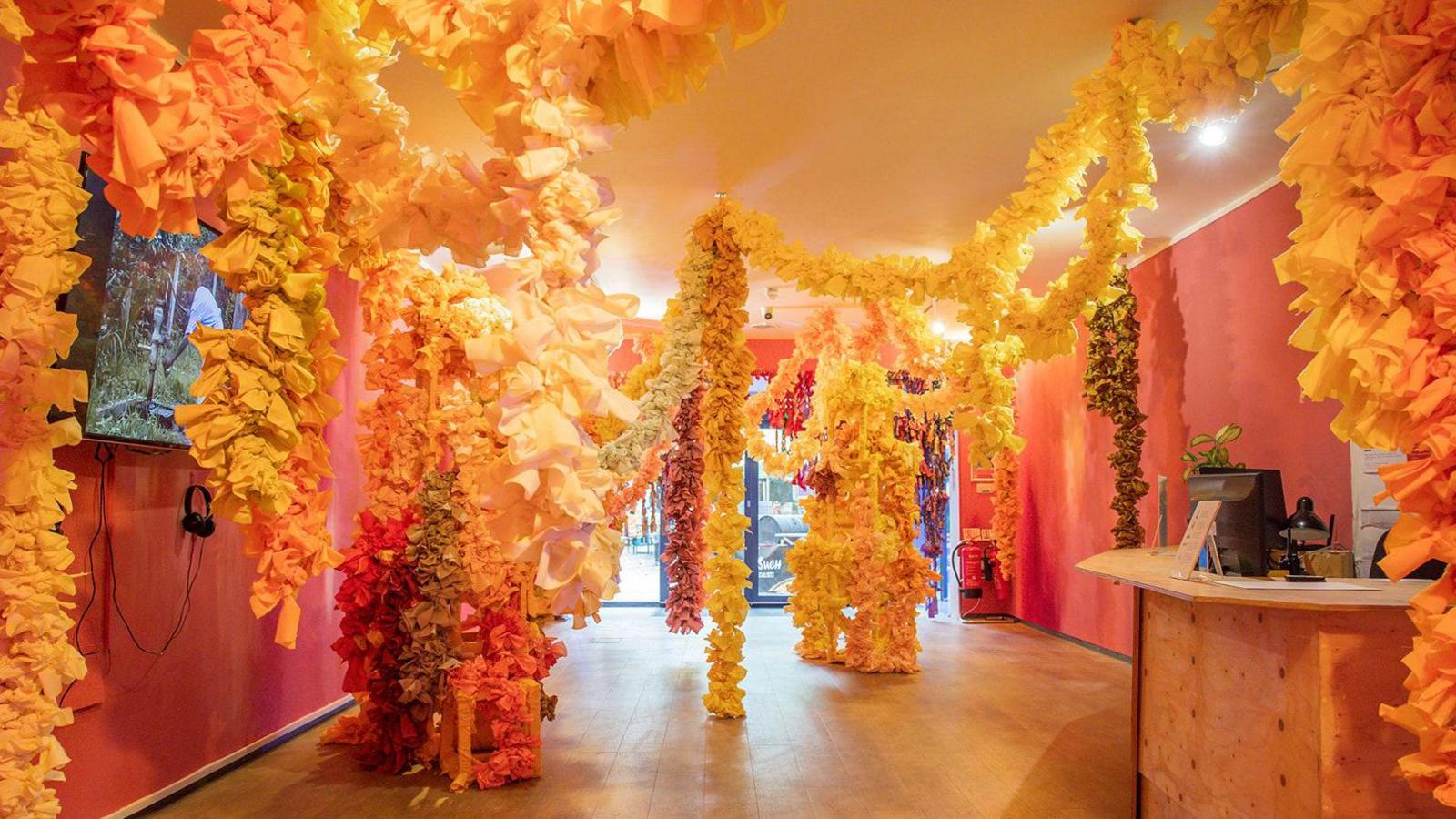 Part of the exhibition space with textiles of yellow and orange and red hanging from the ceiling in flowery displays