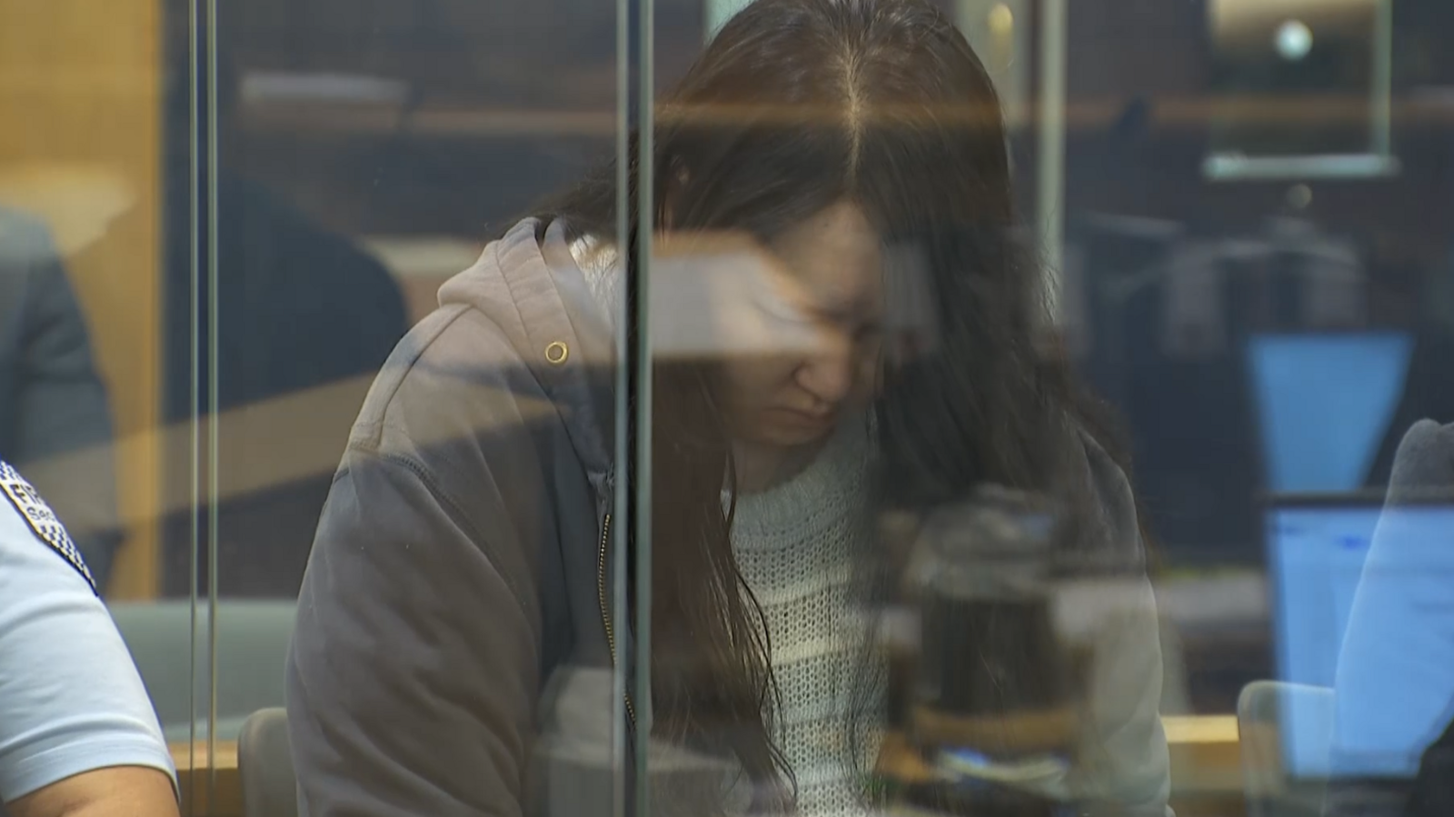 Hakyung Lee, wearing a white sweater and grey hoodie, looks down behind a plastic barrier in the court room