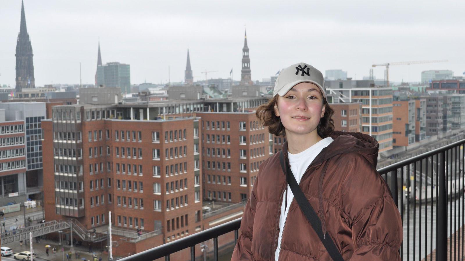 Paulina stands on a balcony in front of a skyline wearing a baseball cap