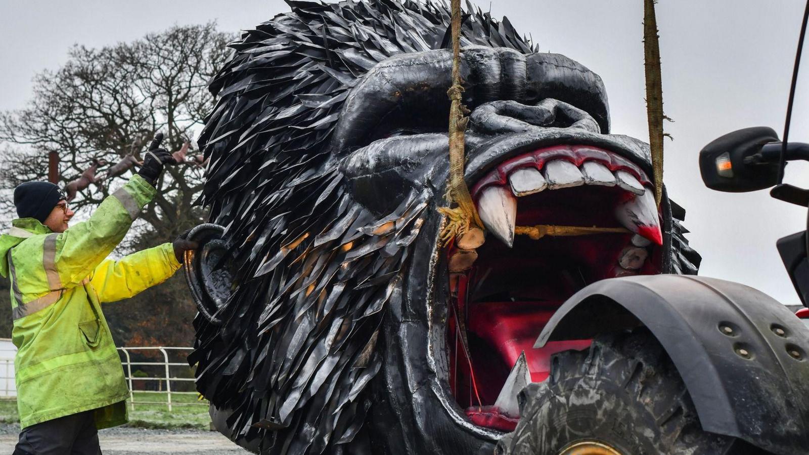 A large roaring gorilla head made of iron, being unloaded off of a machine, attached to ropes 