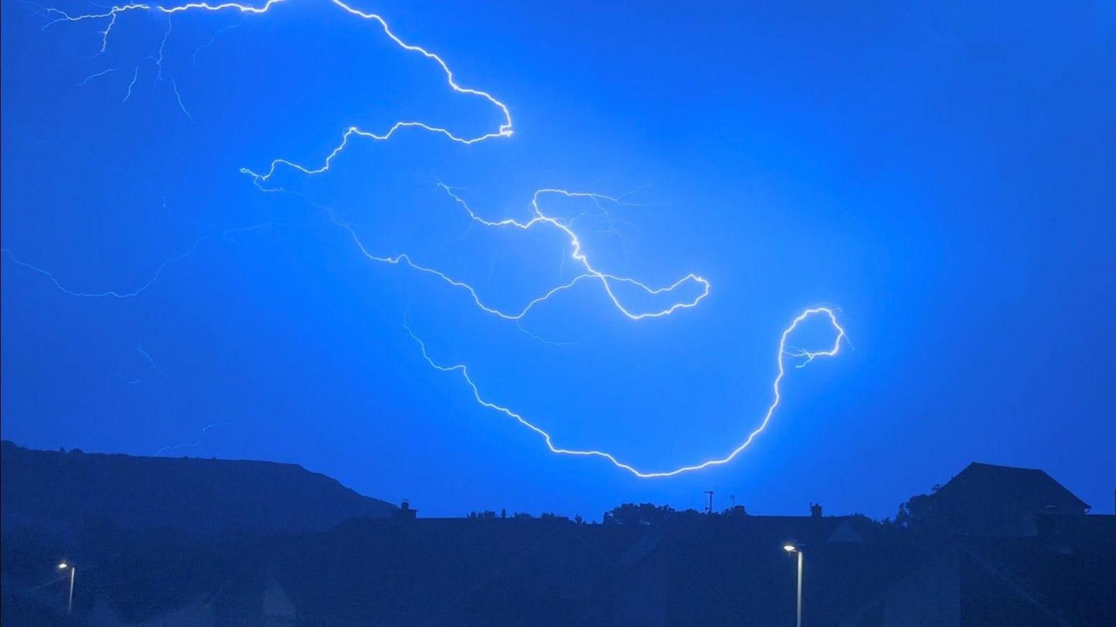 Serveral long lighting bolts across a pale blue night sky with dark hills and houses on the horizon