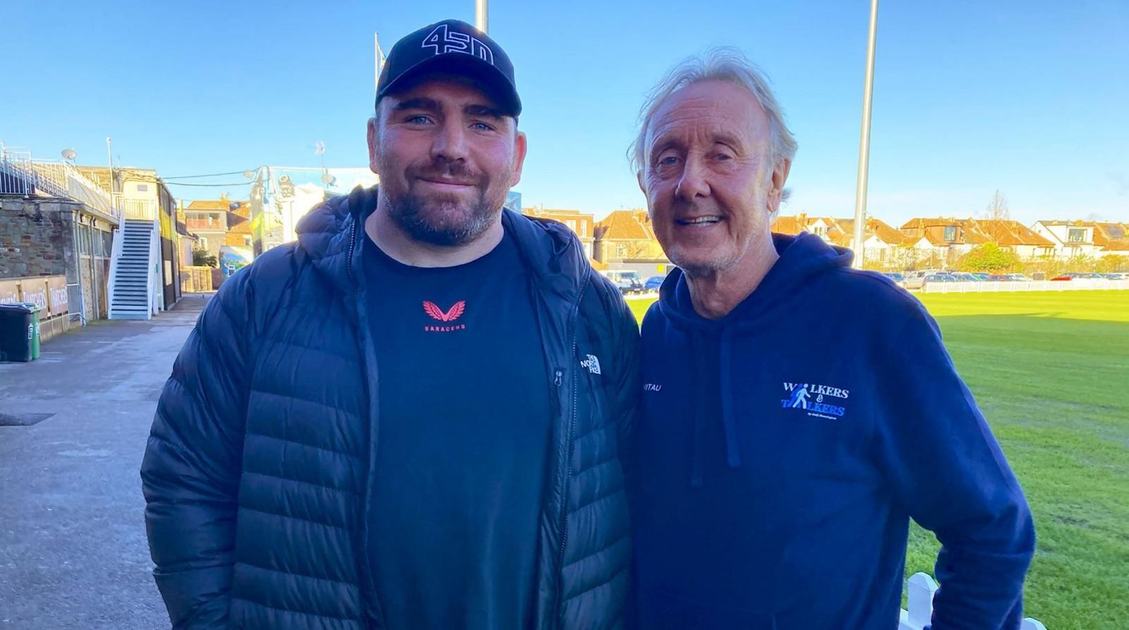 Fraser Balmain with Andy Brassington standing outside a cricket ground in Bristol. They have their arms around each other and both are wearing blue outfits. 