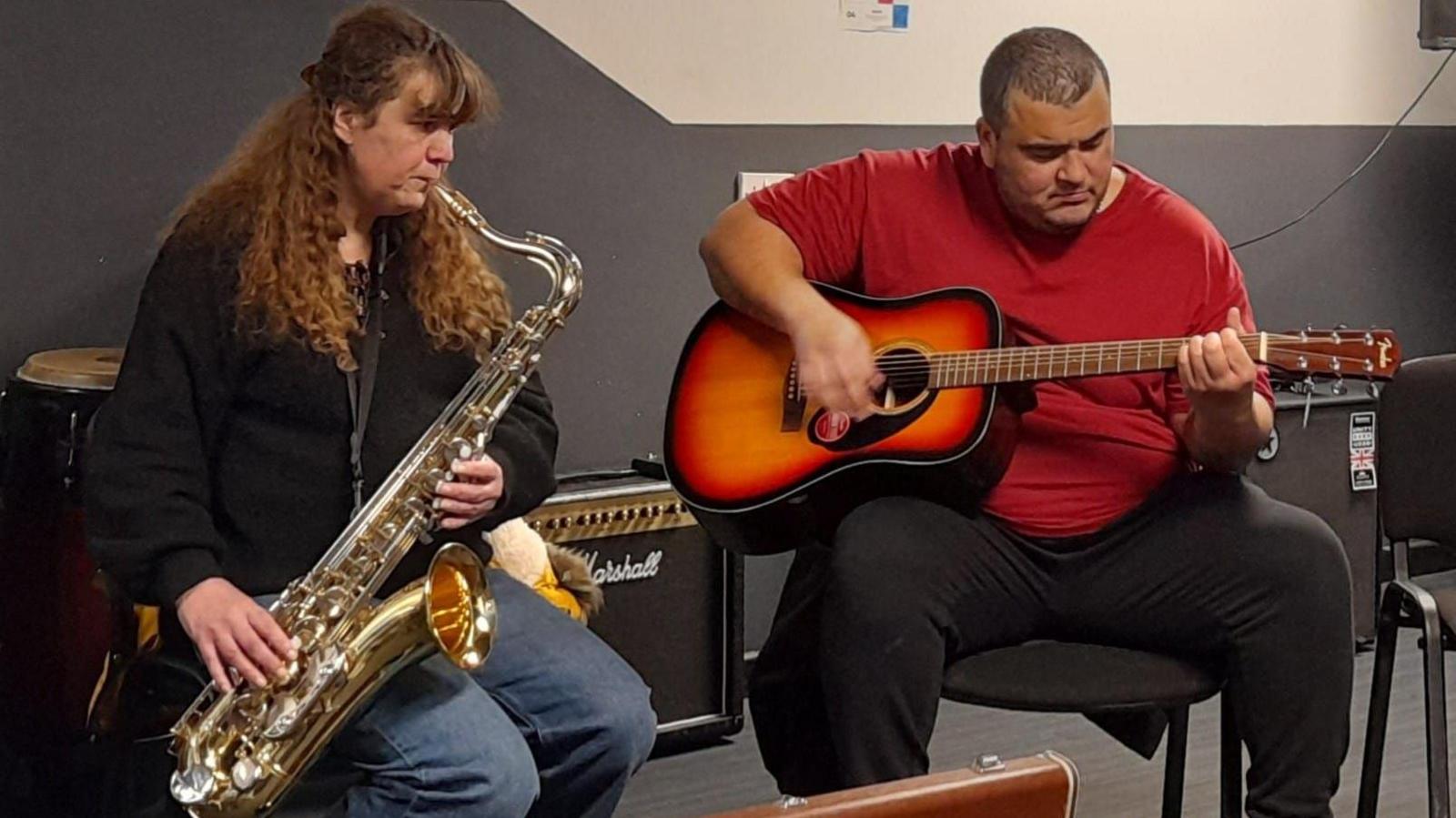 Gaynor sits to the left, she is wearing a black long sleeved jumper and jeans, holding her saxophone as she plays music. To her right is a man with shaved black hair, wearing a short sleeved red t shirt and black tracksuit bottoms playing the guitar.