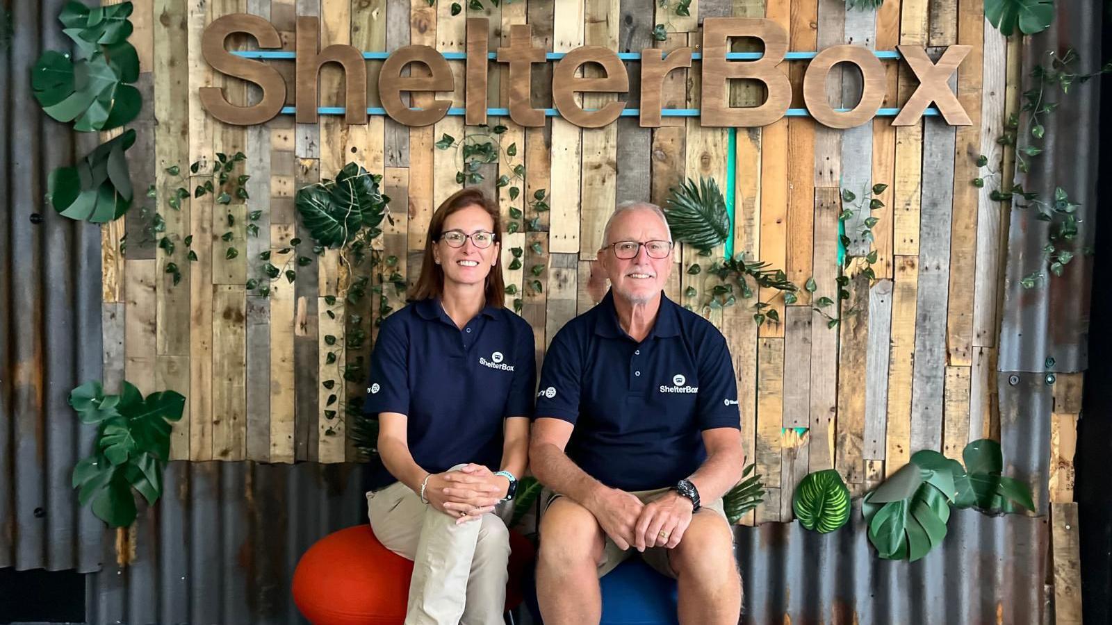 A man and a woman wearing ShelterBox uniforms sit in front of a wooden ShelterBox sign. They are smiling at the camera.