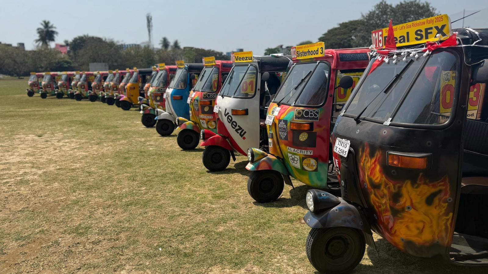 A row of about 17 colourfully decorated and painted tuk-tuks on a mown lawn in India. There are trees in the background under a bright blue sky.