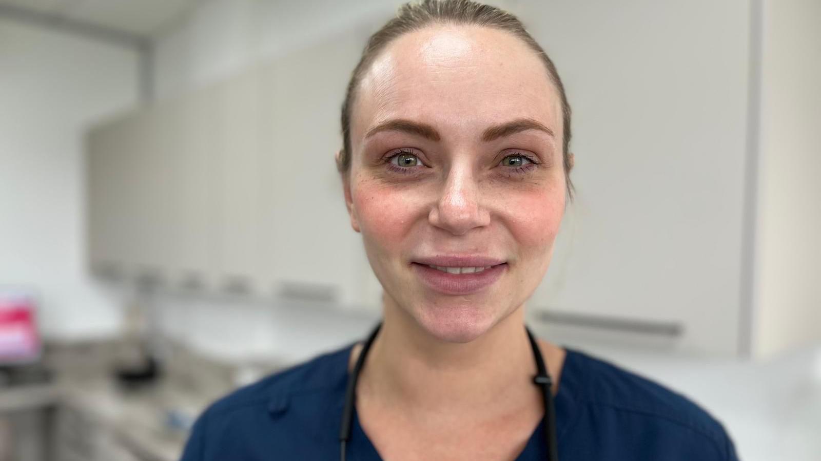 Dr Bassett is standing in a dentist's surgery wearing a navy blue top with her glasses' chain visible around her neck. Her hair is tied back off her face and she is smiling at the camera.