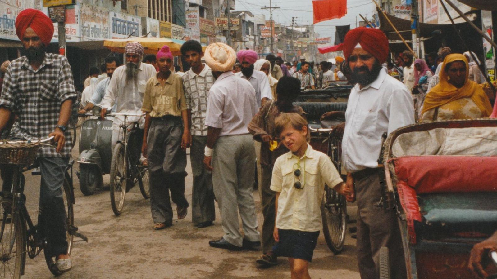 A young boy wearing a white shirt and shorts. He is about six years old and has sunglasses attached to his shirt.
He is holding the hand of his Grandad's who is wearing a red turban and has a black beard. 
He is wearing a light blue shirt and grey trousers with a black belt. They are in India with a lot of people in the background and people riding bikes 
