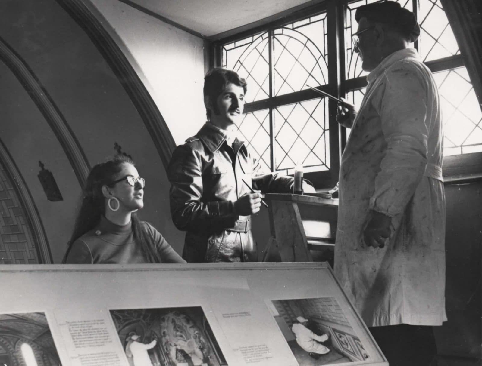 A man in a dirty white coat stands next to a leaded window, holding a paintbrush. He is speaking to two teenagers/ young adults. The girl has long hair, glasses and hooped earrings, the boy is wearing a shirt with a large collar and holds a paintbrush. 