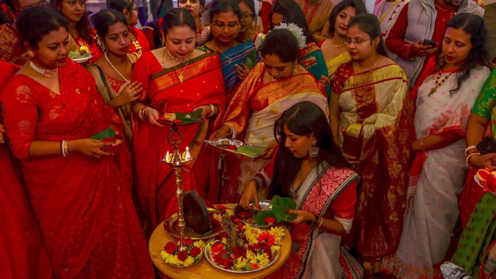 Group of ladies - dressed in indian colourful outfits standing around a candle and trays of flowers 