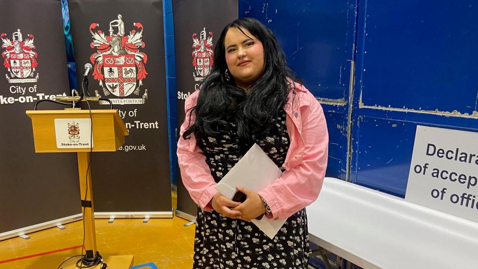 A woman with long black hair, wearing a black dress with white floral pattern and pink jacket. She is standing in a hall during an election count, holding a white folder and iphone. A hoarding in the background says City of Stoke-on-Trent council/
