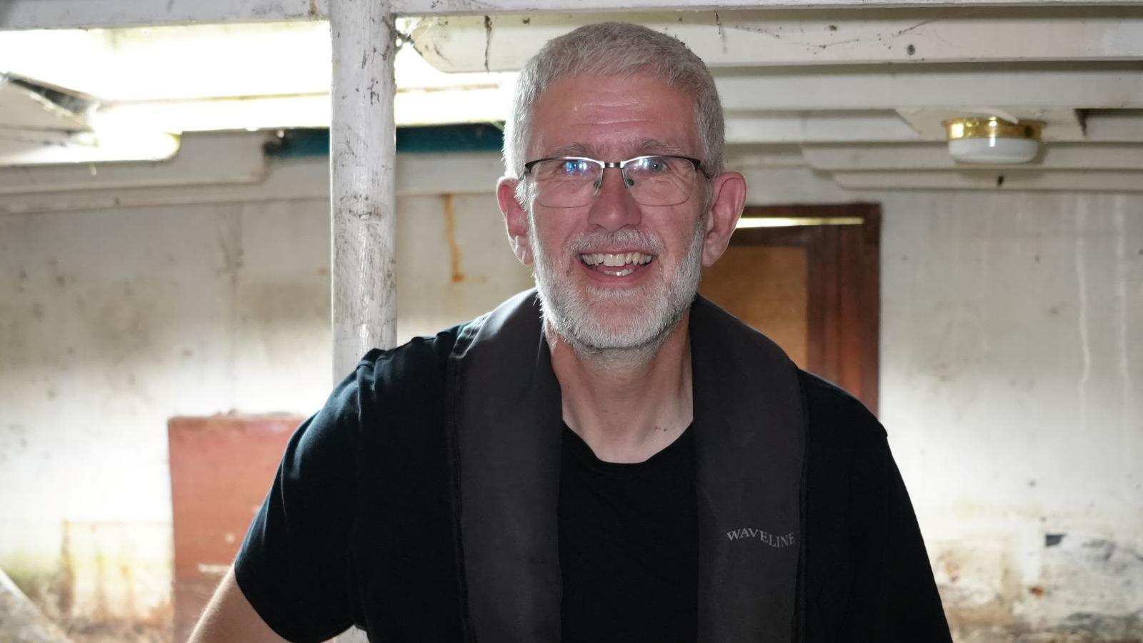 A smiling Tim Shreeve is below deck of the whaleback. The room is decaying and walls are watermarked. He wears a black T-shirt and life jacket.