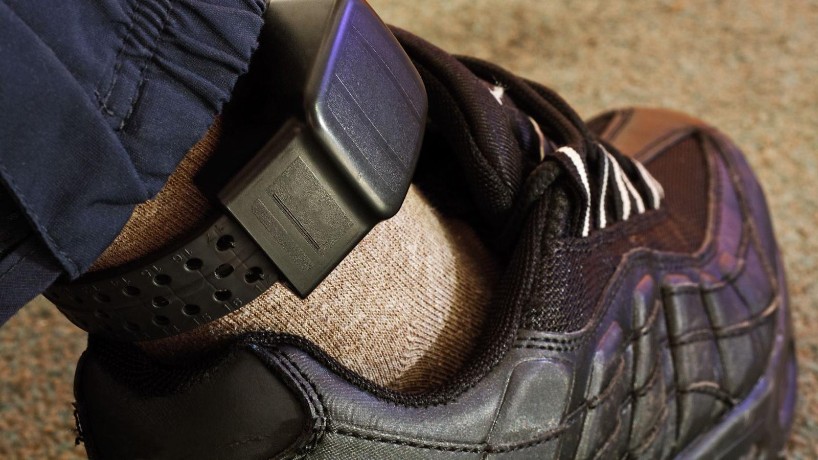 An electronic tag around an ankle. The person wears a black training shoe and light brown sock 