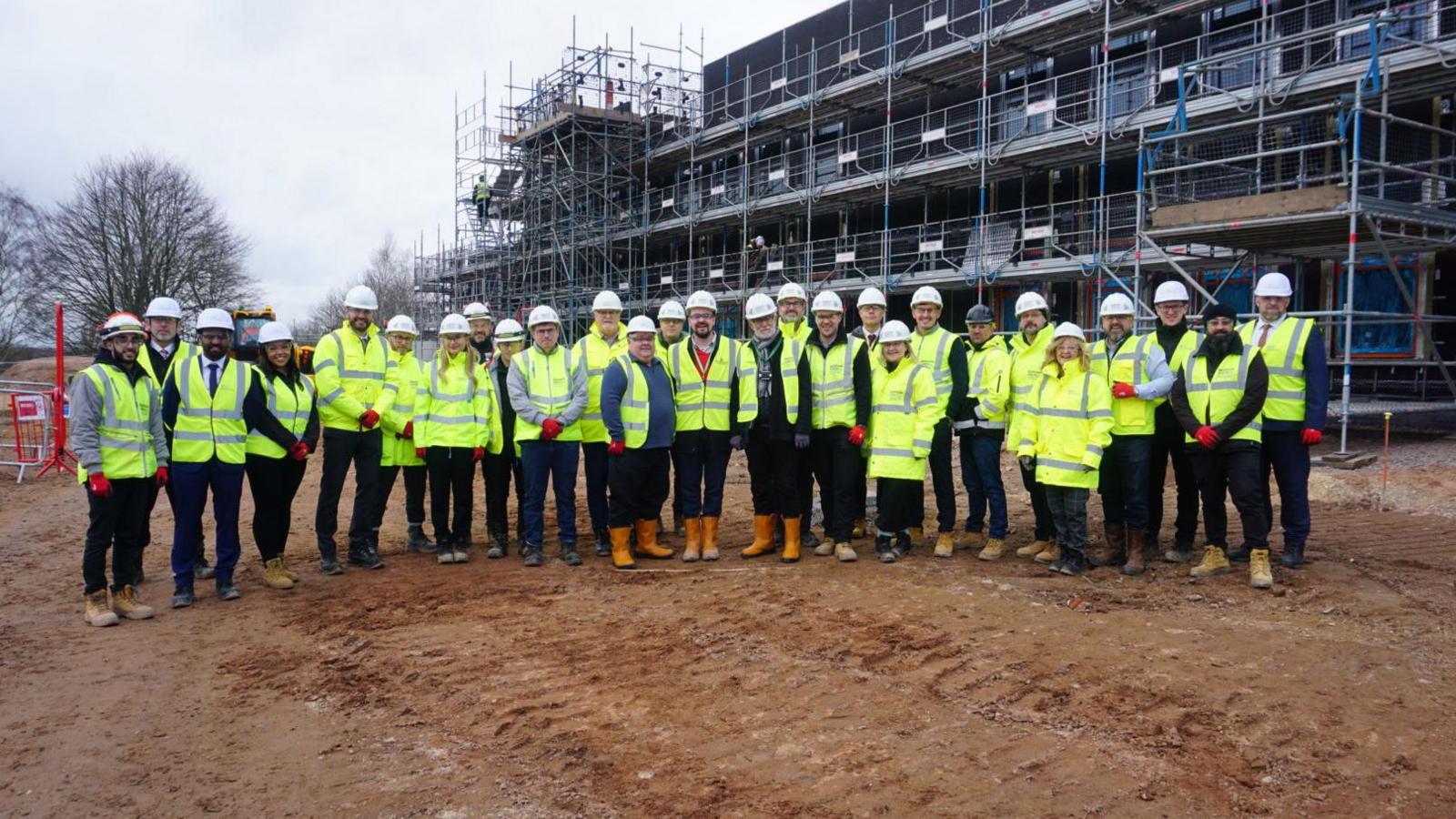 A long line of people wearing high-vis yellow coats and construction helmets.  They stand in front of a large building covered in scaffolding.
