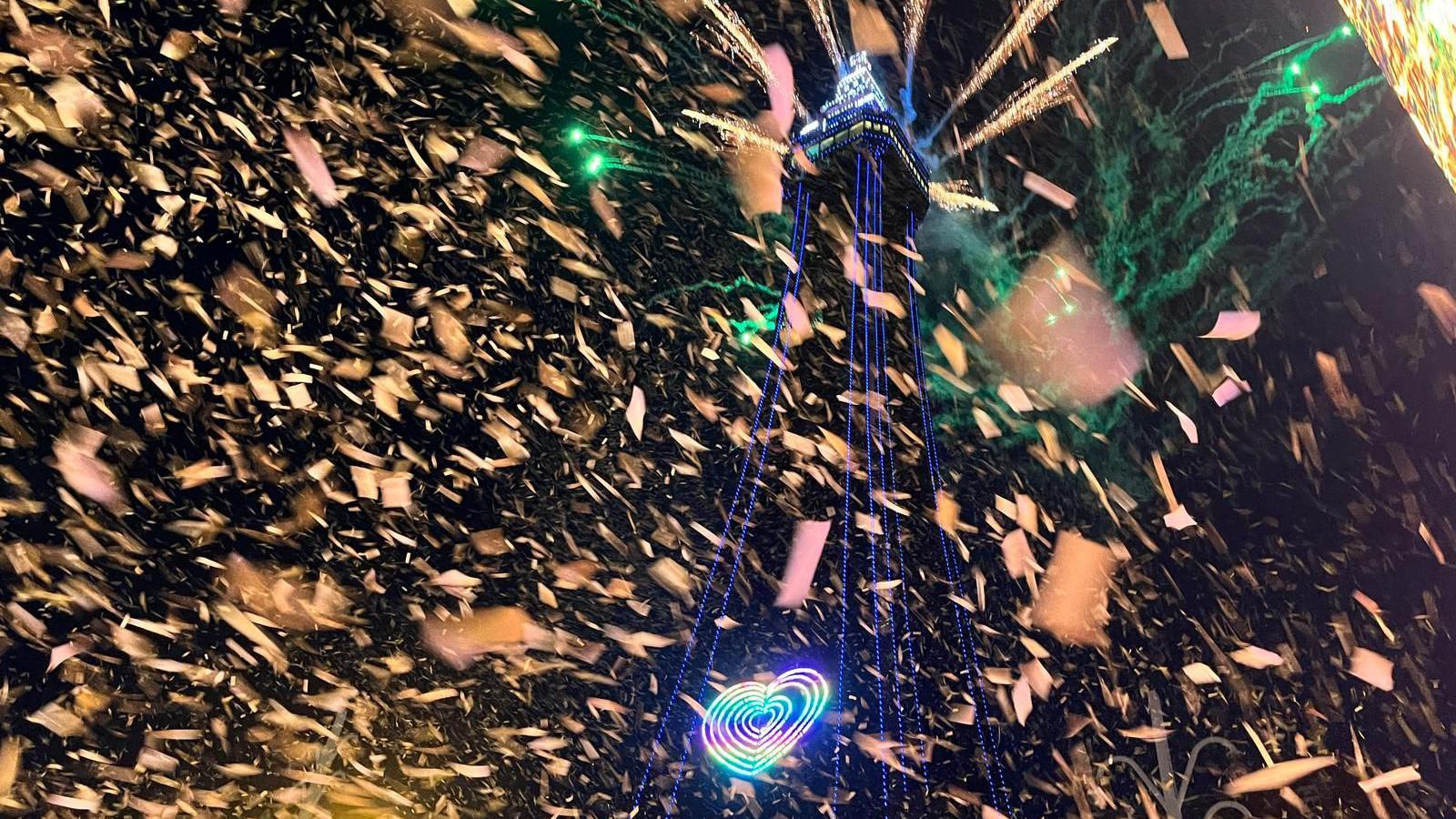 Blackpool Tower is lit up with blue lights and a rainbow-coloured heart and ticker tape. Green fireworks can be seen behind it.