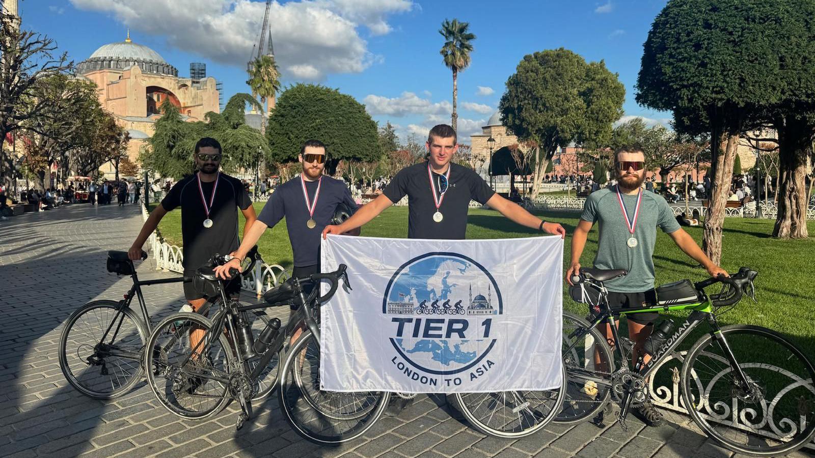 Four men, standing with their bikes, are outdoors with the Blue Mosque in the bcakground. One of the men is holding a white flag with a logo on it that reads Tier 1 London to Asia.
