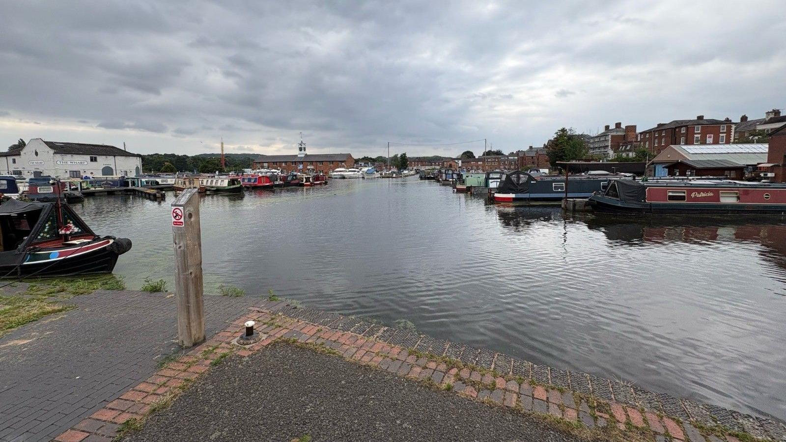 Stourport Basin cleared from duckweed. There is an expanse of water, surrounded on all sides by docked canal boats. There are buildings on either side of the basin.
