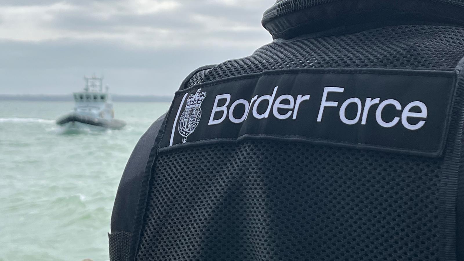 Under grey skies, an anonymous uniformed officer from Border Force looks out to sea with a Border Force vessel in the distance. 