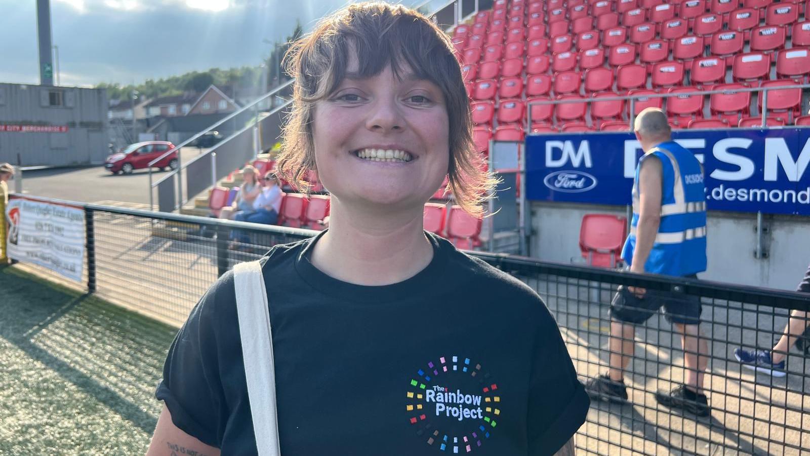 Eimear Willis is wearing a dark T-shirt with the Rainbow Project logo on it. She stands in front of a football stadium stand. A steward in a blue bib walks past her. She has short dark hair and across her shoulder is the white strap of a bag.